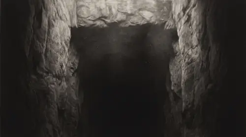 Looking straight down a shaft in bare rock: the walls resolve for six or eight feet in the raking light and then simply stop - no floor, no sound of a floor, the dark going on with no boundary…